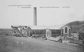 Photo noir et blanc montrant un long bâtiment métallique surmonté d'une cheminée.