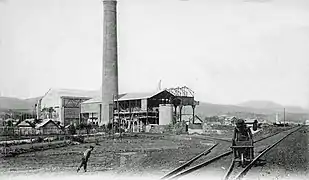 Photo noir et blanc montrant deux bâtiments et une cheminée, une personne pousse une brouette entre des rails.