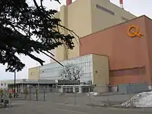 Des portes vitrées à l'entrée d'un bâtiment de couleur brune. Un logo jaune formé d'un Q et d'un éclair orne la façade.