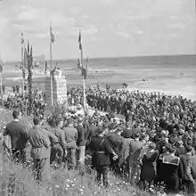 Foule de civils et militaires rassemblés en 1945 à Saint-Laurent-sur-Mer, autour du monument à la mémoire des soldats américains érigé devant la plage d'Omaha Beach, lors d'une cérémonie commémorative marquant le premier anniversaire du jour J.