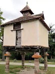 Photo couleur d'un bâtiment carré à structure en bois sur des piliers de pierre. La toiture à 4 pentes en tuiles plates et surmontée en son sommet d'un clocheton en bois percé de trous ronds.