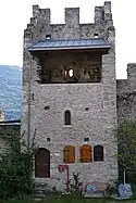 Tour au centre d'un mur en pierre. En bas à gauche sur la tour se trouve une porte puis, à sa droite, deux petits volets en bois. Au milieu du mur de la tour se trouve une petite fenêtre et au sommet du mur, un balcon est protégé par un toit. Le sommet de la tour est crénelé.