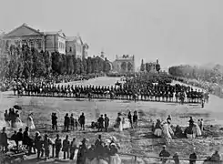 Photo en noir et blanc de militaires assemblés dans une place avec des spectateurs les observant ; en arrière plan, des bâtiments