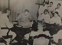 Un groupe de six hommes est assis à une table, l'un d'entre eux signe un document ; ils sont observés par trois hommes debout sur la gauche.