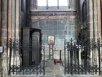 Pièce aux murs partiellement peints et accessible par une grille en fer forgé ouverte. À l'intérieur, un confessionnal, une icône accrochée au mur, un prie-Dieu et un autel