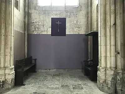 Pièce contenant un banc et un siège en bois. Les parties basses des murs sont peintes en violet. Un crucifix est accroché au mur.