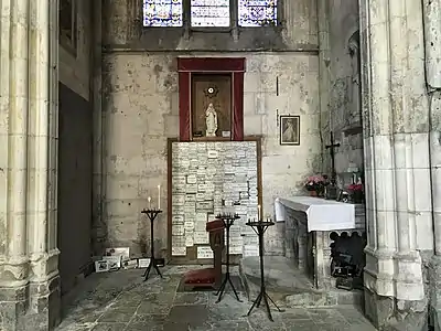 Pièce avec des murs de pierre, contenant un prie-Dieu, des repose-cierges et un autel. Sur le mur face au photographe, de nombreux ex-voto sont surmontés par une statue de femme dans une niche.