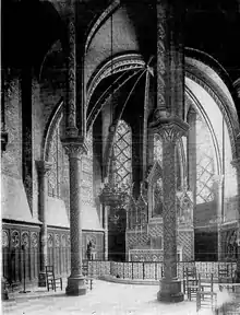Photographie en noir et blanc. Au premier plan, deux piliers supportent une voûte. En arrière-plan, peu visible, un autel richement décoré.