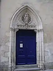 Deux marches d’escalier, une porte bleue à deux battants, un tympan en ogive décoré finement, avec une statue de la Vierge devant une rosace, et deux bas-reliefs d’anges.