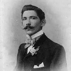 Un homme à moustaches pose dans un costume avec noeud papillon.