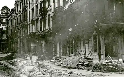 Photo ancienne en noir et blanc d'une rue avec des bâtiments ruinés. Des débris jonchent la chaussée.