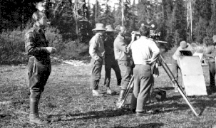 Photographie en noir et blanc d'un groupe d'hommes rassemblés devant une caméra sur pied, en extérieur ; personnage principal à l'écart.