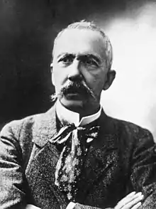 Photo en buste d'un homme en costume se croisant les bras et portant une moustache.