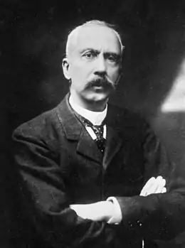 Portrait en noir et blanc d'un homme moustachu portant un costume et croisant les bras.