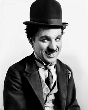 Plan poitrine d'un homme au chapeau rond, petite moustache noire, en costume élimé qui regarde de côté d'un air rieur