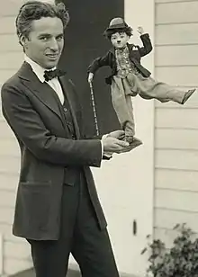 Photographie de Charlie Chaplin debout et en costume tenant une poupée représentant Charlot