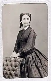 Charlotte photographiée en noir et blanc a l'air mélancolique, elle se tient debout, les mains posées sur le dossier d'un fauteuil capitonné