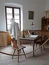 Photographie d’un moine debout dans une cellule comprenant une table, une chaise et des livres.