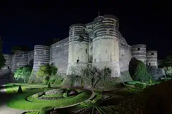  Photographie d'un château éclairé la nuit, six tours imposantes et des jardins au premier-plan dans les douves.