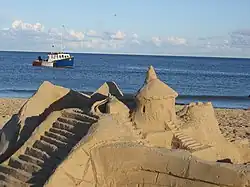 Photographie d'un château de sable sur une plage des îles de la Madeleine. Un bateau est visible au second plan, peint comme le drapeau de l'Acadie.