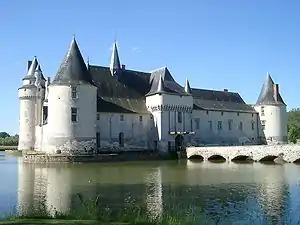Photographie couleur de loin et de côté d'un château médiéval de type forteresse féodale en pierre blanche, situé au milieu d'une pièce d'eau, et du pont qui y mène.