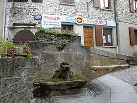 Photographie montrant une fontaine en pierre devant des maisons également en pierre.