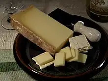 La photographie en couleur représente une tranche de beaufort dans une assiette brune, posée sur une nappe à gros grain. Dans l'assiette se trouvent aussi des tranchettes de fromage, des crackers et un couteau à fromage large en inox à manche en marbre ; un exemple de service du beaufort à l'apéritif