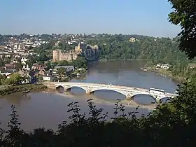 Vue ensoleillée sur un pont métallique blanc en contrebas séparant les deux rives d'un large fleuve. Sur la rive de gauche sont visibles les contours d'une grande ville avec un château médiéval, et sur la rive droite une zone plus rurale