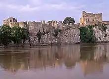 .Château de vielles pierres en ruine, formé de murs bruts et de larges tours, s'étalant en longueur au sommet d'une petite falaise, au pied de laquelle coule une rivière. Le ciel est bleu pastel et le château est rosé, dû à l'heure matinale à laquelle la photo a été prise.