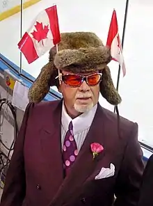  Photographie couleur de Don Cherry en costume-cravate dans les tribunes d'une patinoire. Il porte une chapka surmontée de deux drapeaux canadiens.