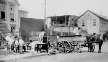 Photo en noir et blanc, une charrette à cheval dans la rue est chargée par un homme tandis que sa famille regarde