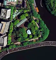 Photo couleur : vue aérienne d'une étendue boisée, d'où émergent quelques bâtiments (l'un d'eux présente une toiture de forme hexagonale), au bord d'un cours d'eau.