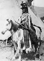 Photographie noir et blanc d'un Amérindien monté sur un cheval blanc, armé d'un arc et coiffé d'un chapeau. Une tente indienne est partiellement visible en arrière-plan.