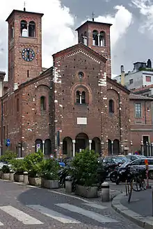 Photographie en couleurs d'une église en briques rouges trapue, avec au premier plan un passage clouté, des bacs à plantes, un parking à voitures.