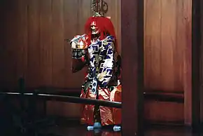 Acteur debout en habit traditionnel bariolé, masque effrayant et grande crinière rouge.