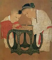 Peinture de deux enfants jouant autour d'une table. Au gauche, un jeune garçon porte un pantalon blanc et une veste rouge. À droite, une fille, plus âgée, est drapée de blanc et porte une ceinture orange.