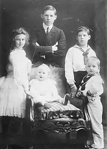 Photographie montrant quatre enfants (une fille et trois garçons) entourant un bébé assis sur une chaise.