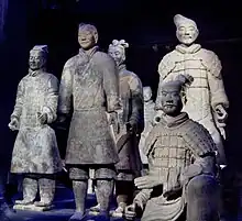Photographie de six statues de soldats en terre cuite coiffés et vêtus distinctement, l'un assis au premier plan et les autres debout.