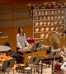  photo de la section percussion d'un orchestre avec une femme jouant d'un instrument au centre et derrière elle, à droite, un yunlao à 37 gongs.