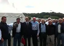 Un homme en costume est entouré d'autres hommes dans un stade de rugby près d'une réception.