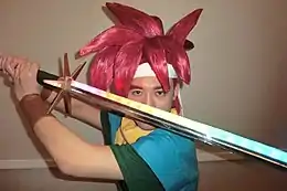 Homme déguisé en Chrono (personnage de Chrono Trigger), aux cheveux rouges montre une épée devant son visage.