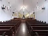Photographie couleur de l'intérieur d'une église sans bas-côté ni transept, entièrement peinte en blanc, avec des bancs de bois.