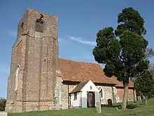 Photo en couleurs d'un long bâtiment dominé par une grande tour carrée à une extrémité, entouré d'une pelouse où apparaissent quelques stèles funéraires