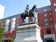 Harrison porte un bicorne et un uniforme militaire. La statue équestre en bronze se trouve un piédestal blanc devant des bâtiments en briques rouges.