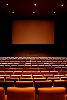 Salle de cinéma, avec plusieurs rangées de fauteuils rouges et un écran géant blanc en fond.