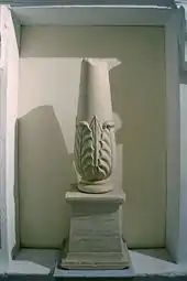 Photographie représentant un cippe, petite colonne tronquée en haut, renflée à la base et décorée d'un motif de feuillage. Elle repose sur un socle trapézoïdal dont la face avant porte les inscriptions - en phénicien au-dessus, en grec en dessous.
