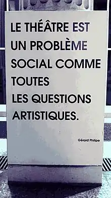 Gros plan sur un panneau rectangulaire à la base d'une colonne où il est écrit en noir sur fond blanc la citation : "Le théâtre est un problème social comme toutes les questions artistiques. Gérard Philipe"