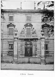 Photo en noir et blanc d'un bâtiment volets clos, avec devant un portail ouvragé fermé.