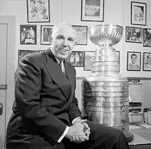 Photographie en noir et blanc de Campbell assis sur le bord d'une table sur laquelle est posée un trophée, la Coupe Stanley
