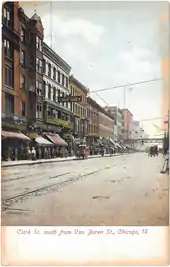 photo colorée d'une large avenus bordée d'immeubles avec beaucoup de boutiques, la foule sur les trottoirs, et deux voitues à cheval.